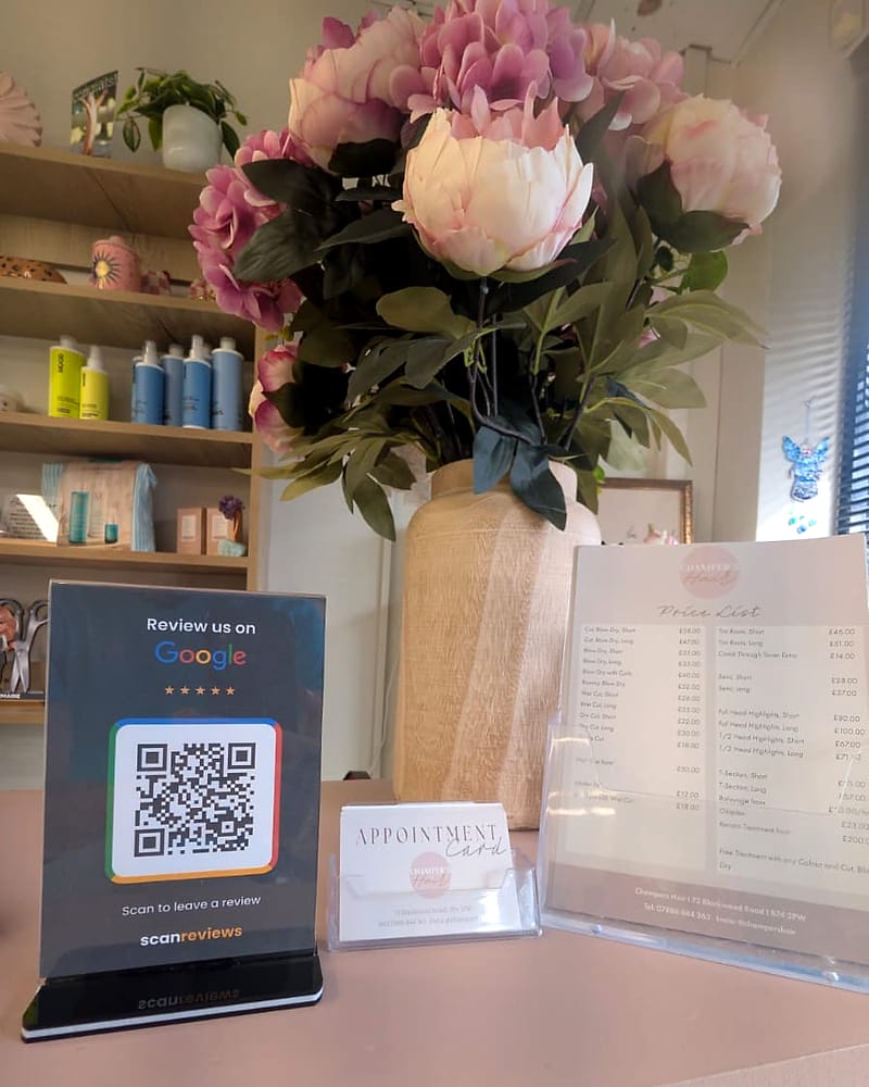 A ScanReviews Google review card in its acrylic stand on a hair salon front desk, beside the appointment cards and price list